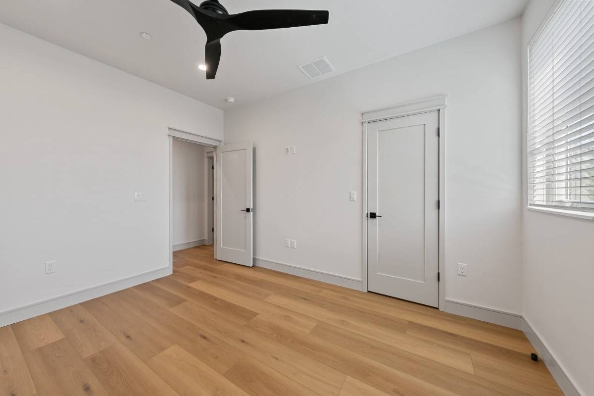 Empty bedroom with light wood flooring, white walls, two closed doors, and a ceiling fan.