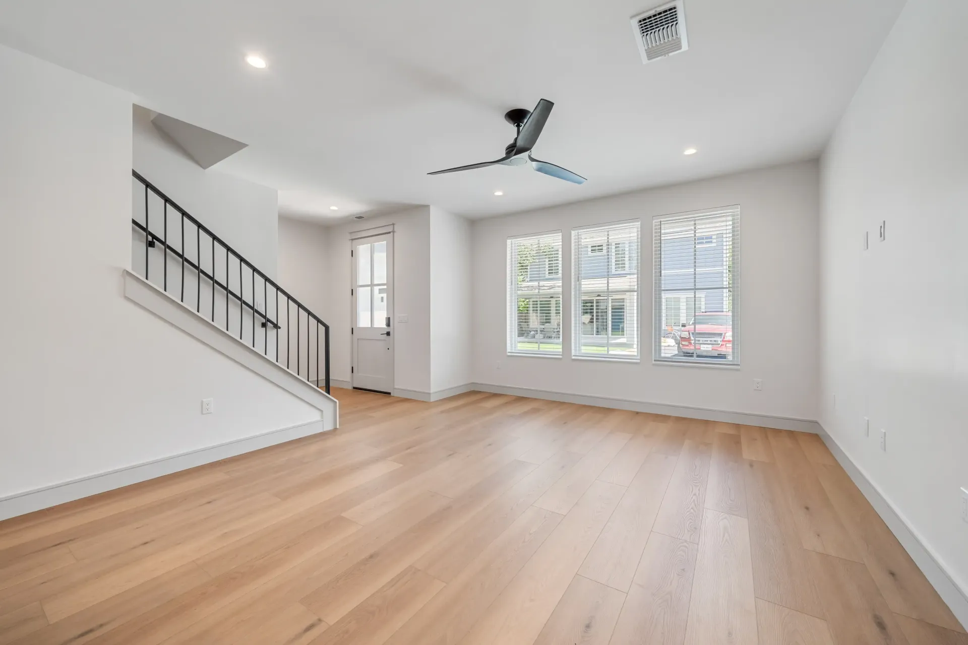 Empty room with light wood floors, white walls, stairs, windows, and ceiling fan.