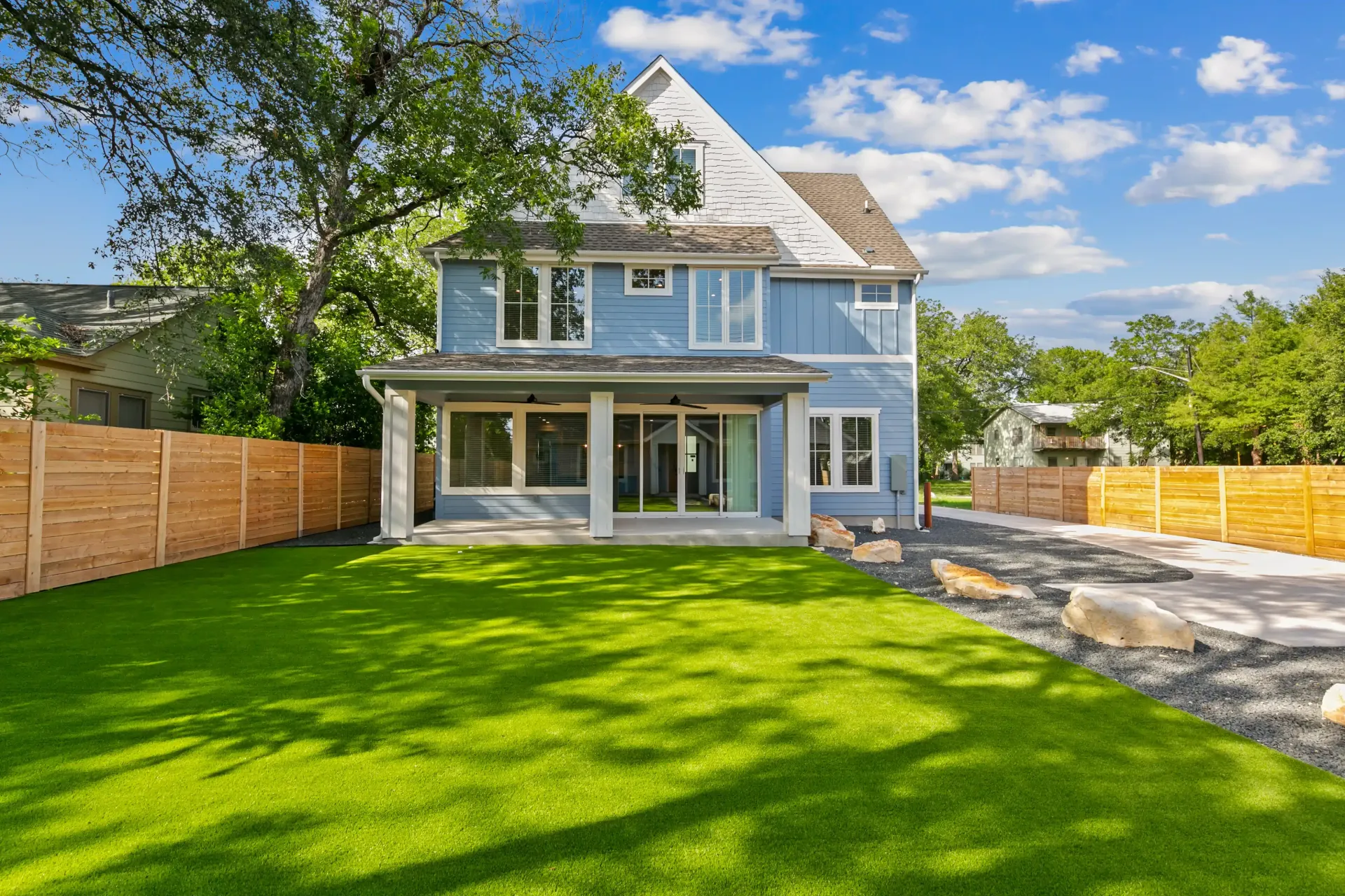Blue two-story house with covered back patio, green lawn, wooden fence, and paved driveway.