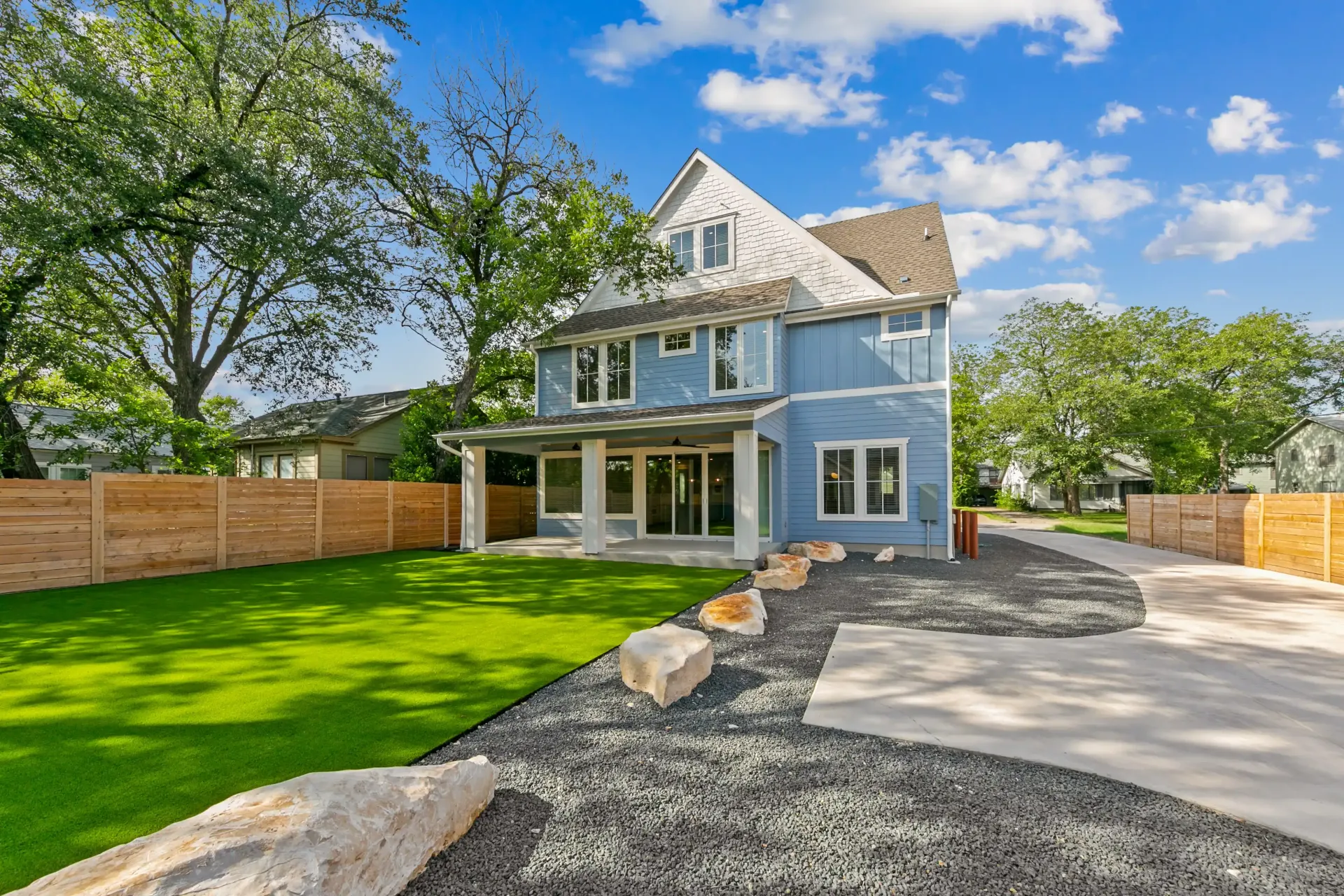 Two-story blue house with porch, gravel walkway, and grassy yard. Wooden fence and trees in background.