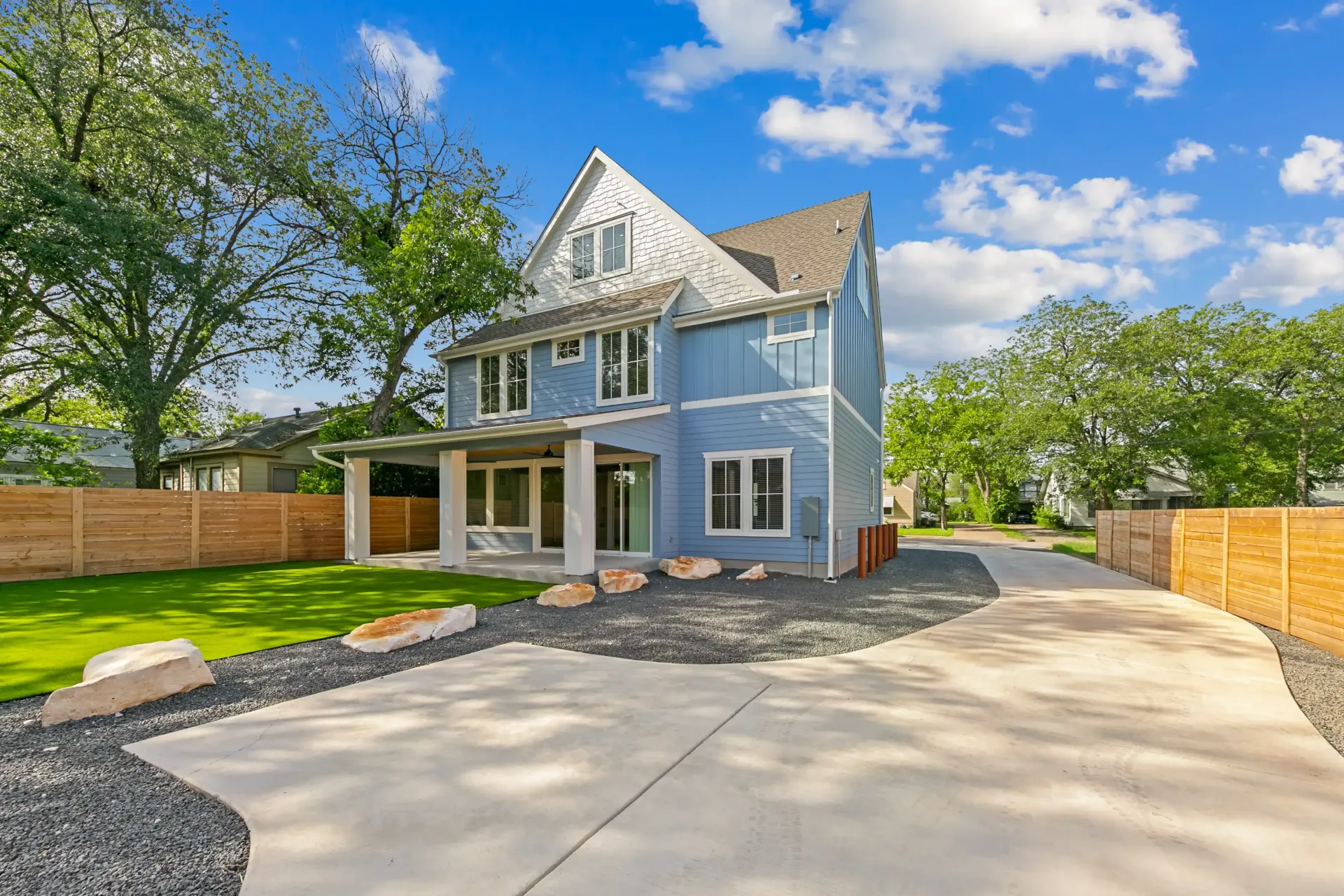 Blue two-story house with covered porch, wooden fence, and concrete driveway. Sunny day with blue sky.