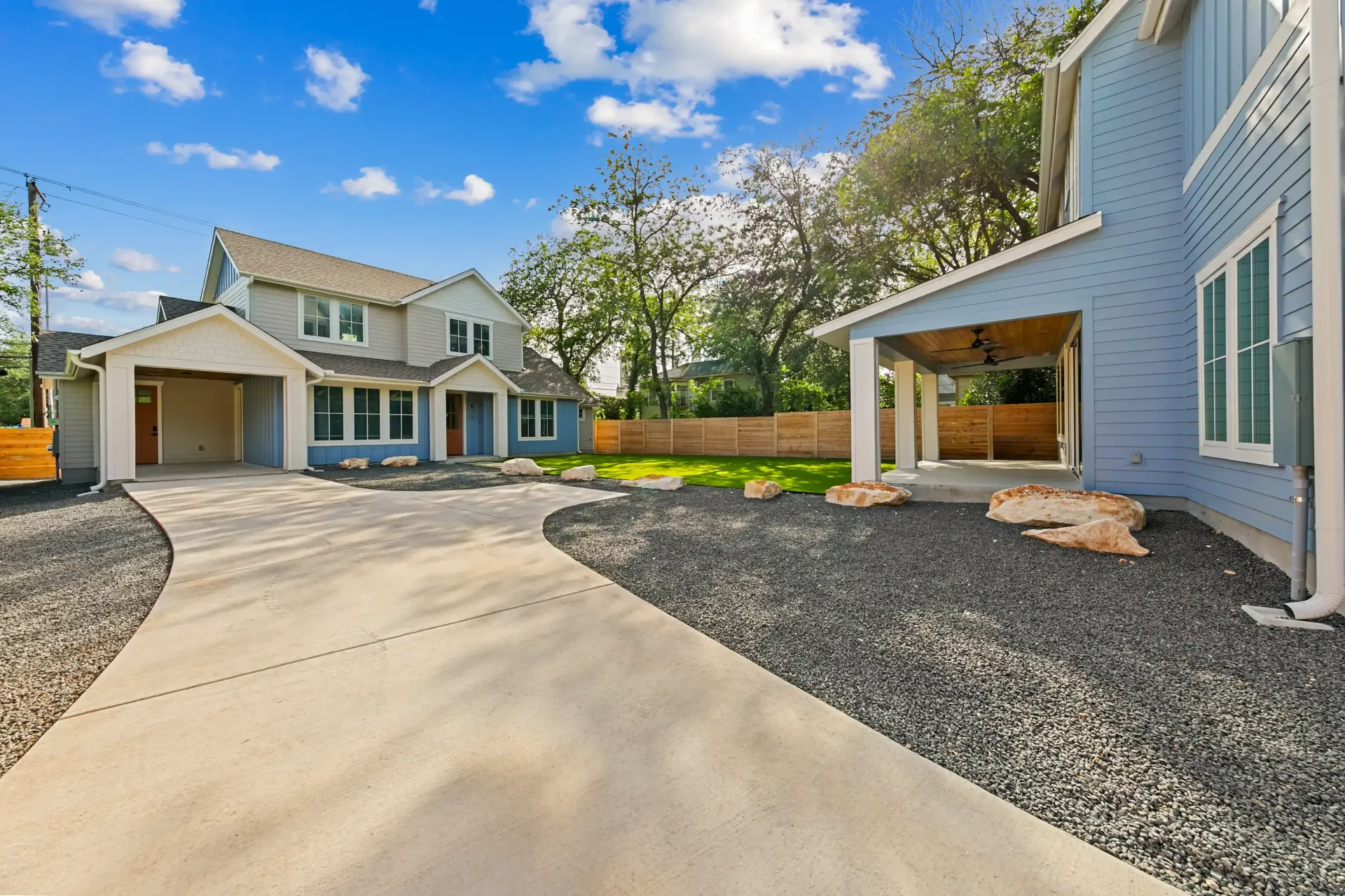 Two-story light blue house with two carports, gray gravel driveway, blue sky.