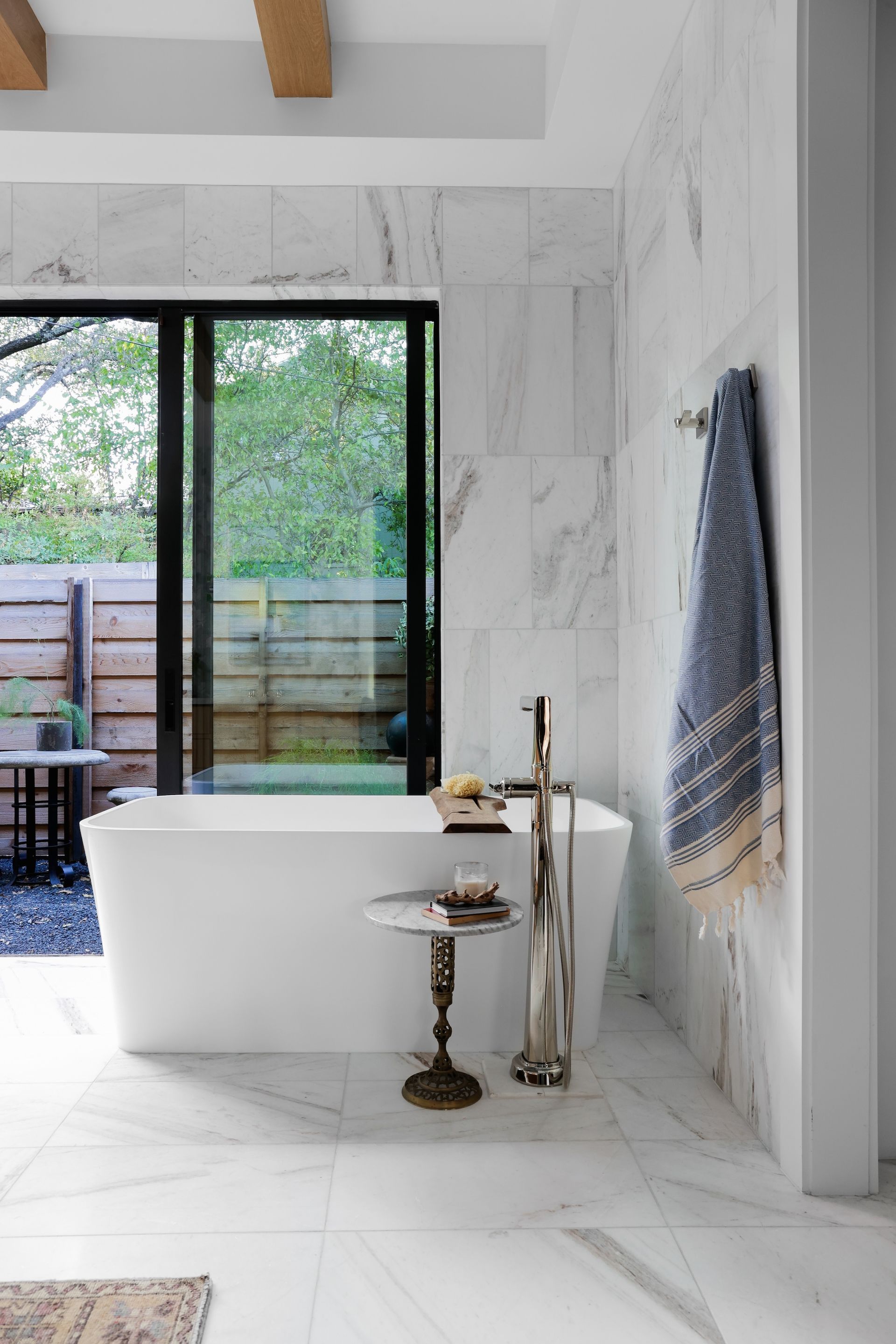 A modern bathroom featuring a white soaking tub next to a large window, a small side table, and a blue towel on the wall.