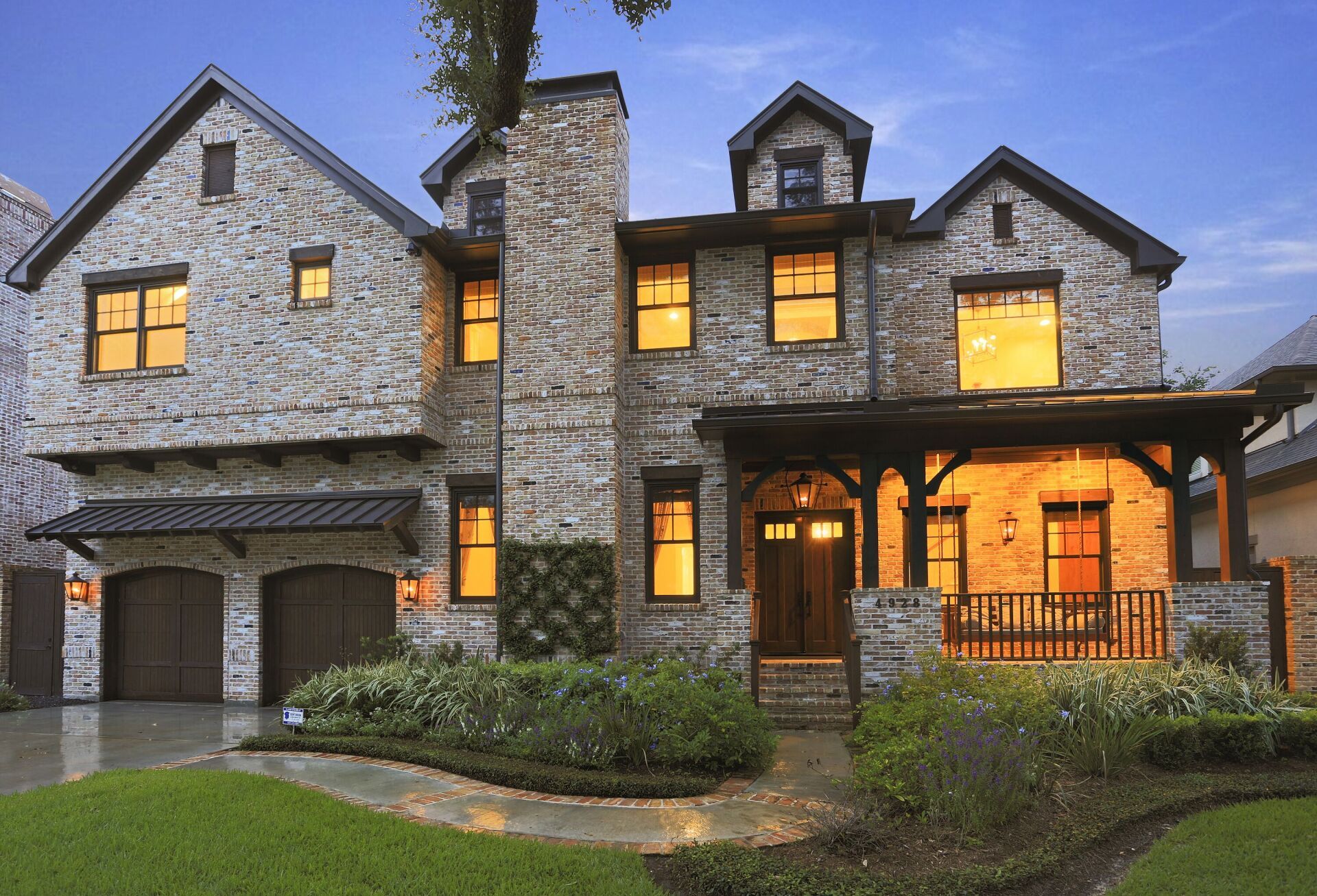 Stone-clad house with a porch and garage, lit from within at dusk. Lush landscaping in front.