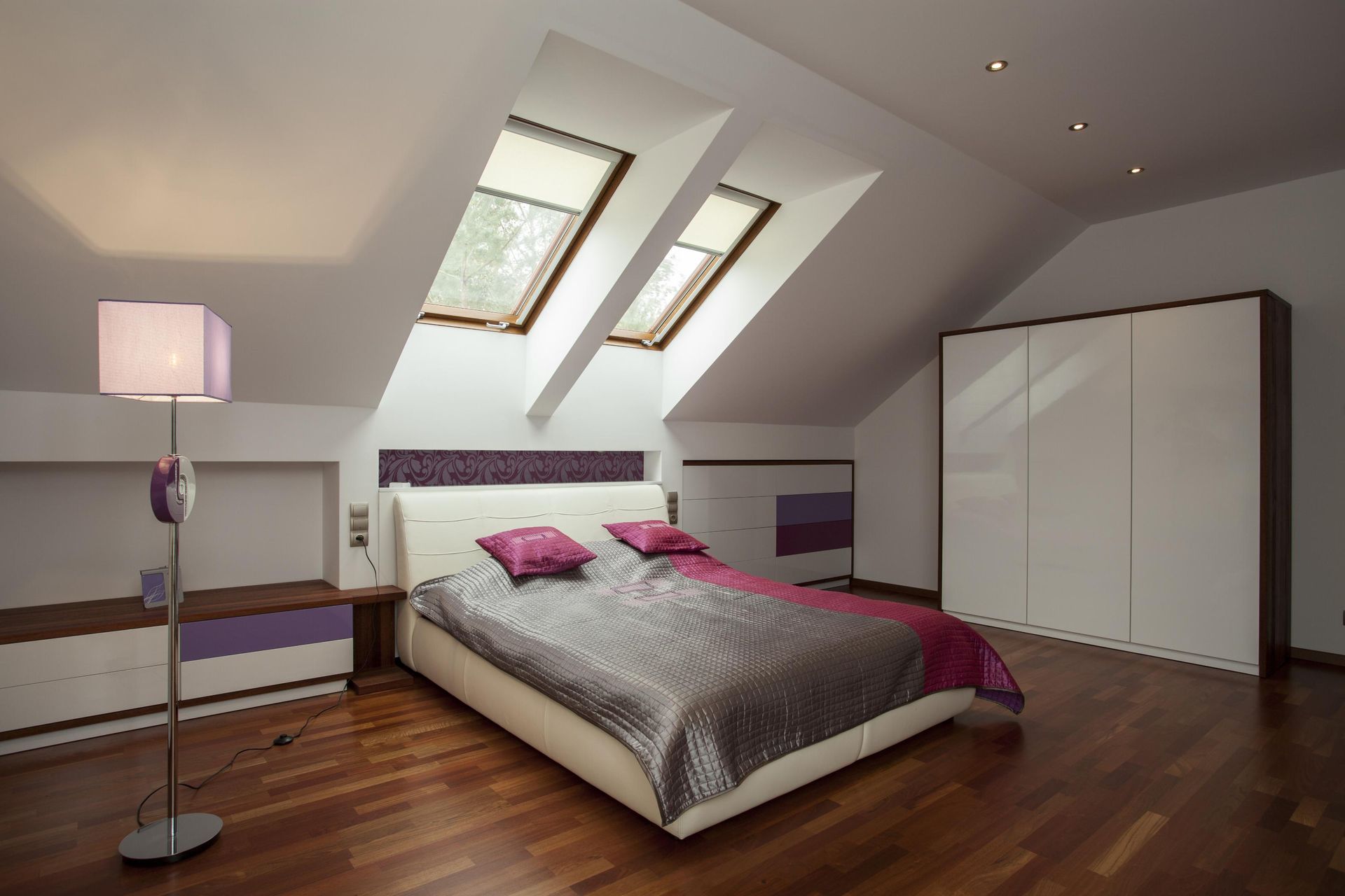 Modern attic bedroom with slanted skylights, wooden floor, bed, floor lamp, and white wardrobe