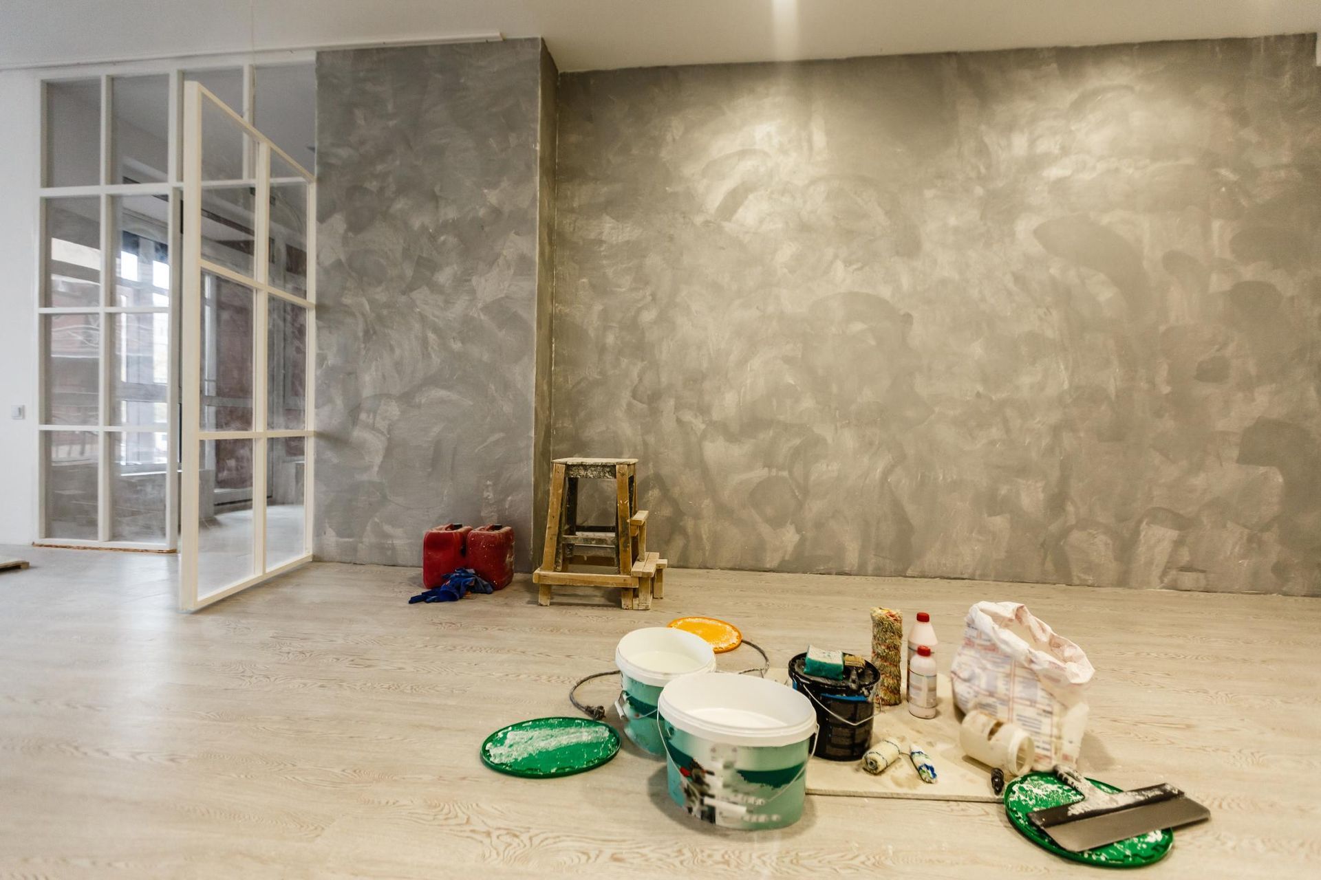 Paint supplies on a floor in a nearly finished room with textured gray walls and white glass doors.