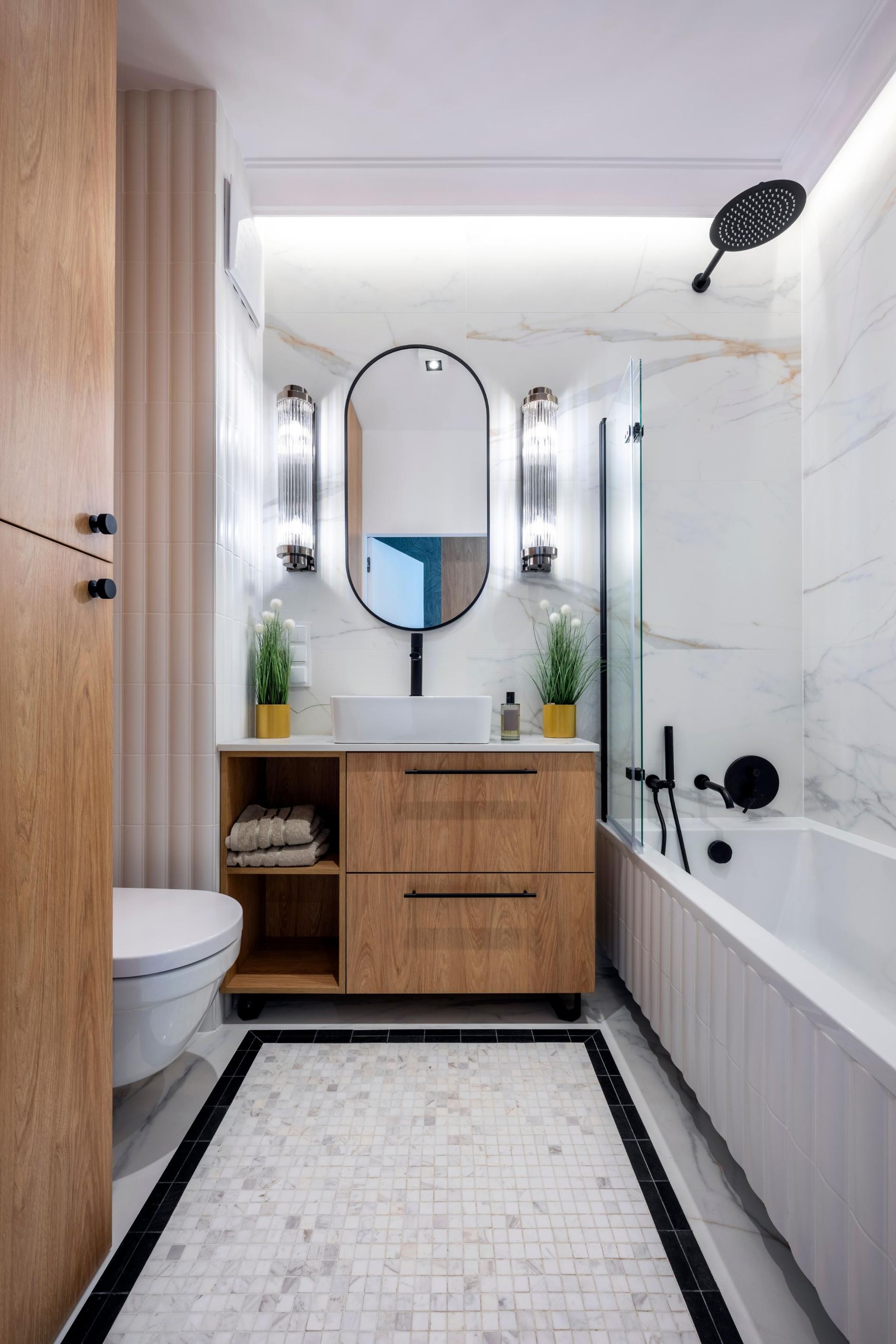 Modern bathroom with white marble walls, black fixtures, wood vanity, oval mirror, and bathtub.