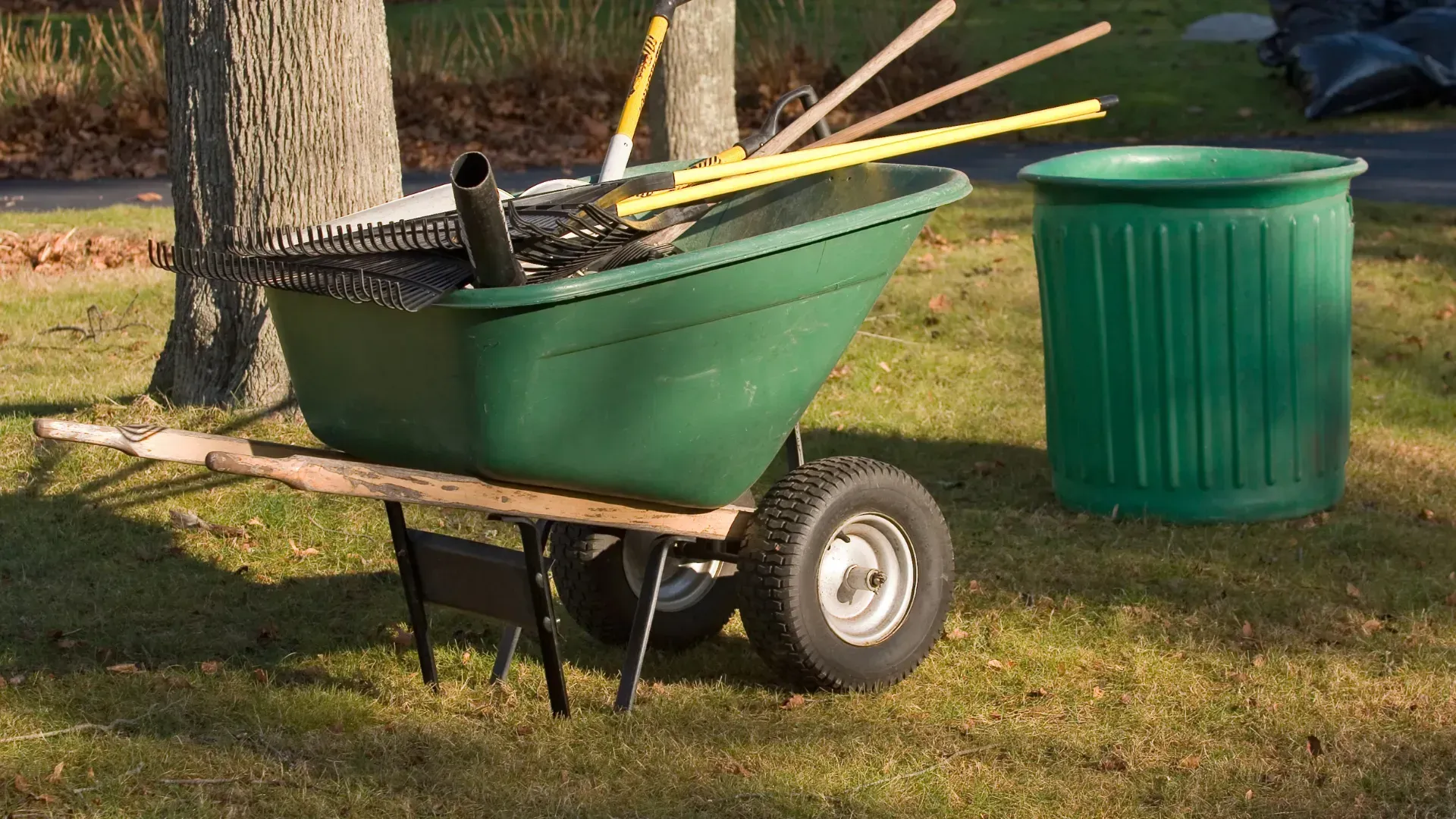 A green wheelbarrow filled with gardening tools next to a green trash can