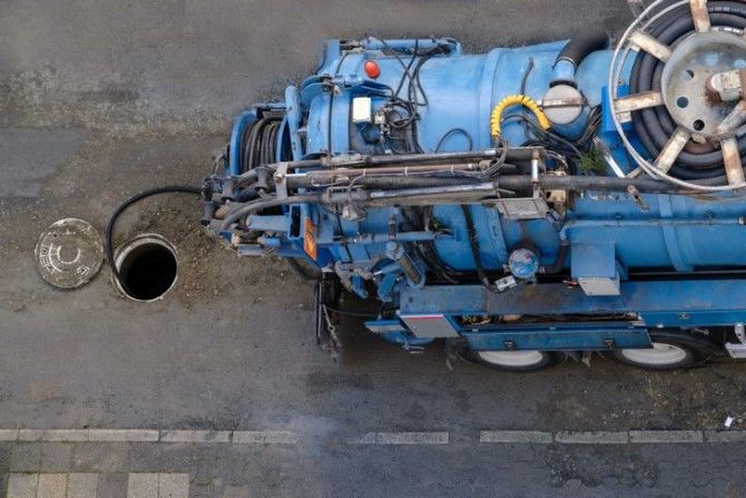 Blue sanitation truck parked over an open manhole, black hose inserted.
