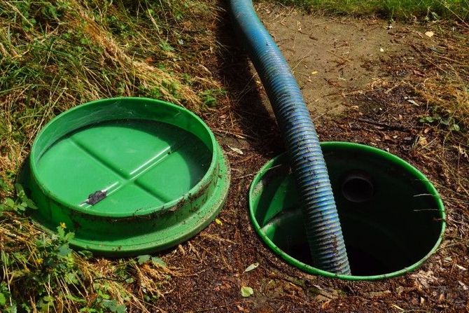 Open green septic tank with hose, cover beside it, in grassy outdoor setting.