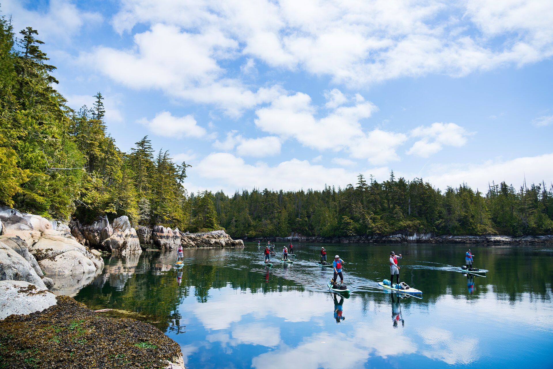 Kayakers on a calm forest lake reflecting blue sky and trees, with rocky shore and dense greenery.