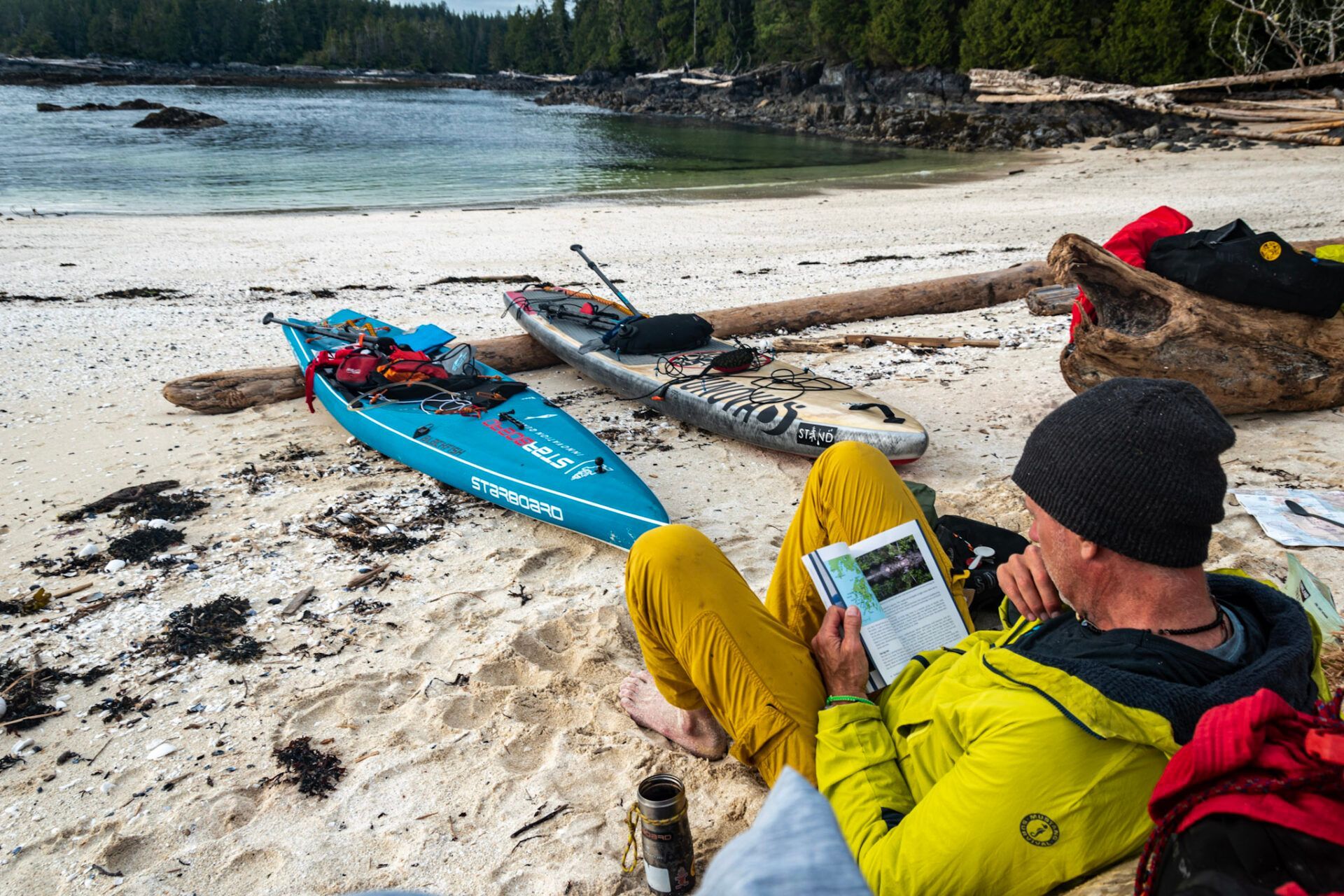 Kayaker in yellow jacket reading on a sandy beach beside a blue kayak and gear near calm water