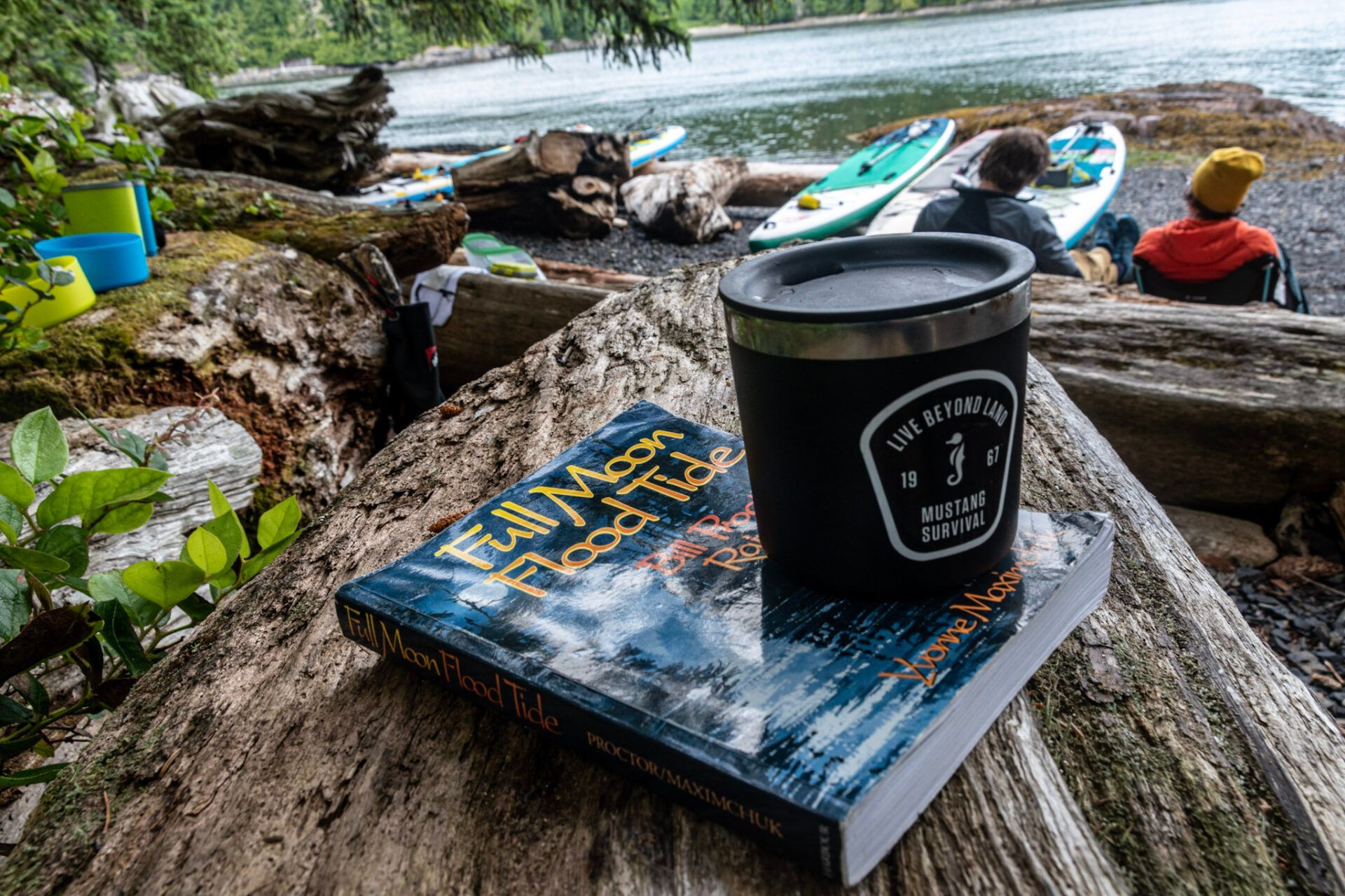 Coffee cup on a book by a lakeside dock with kayaks and a seated person in the background
