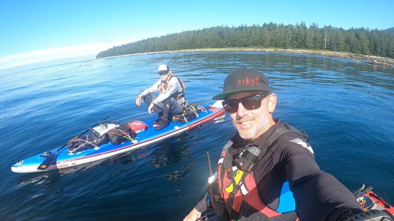 Two kayakers on a calm blue lake near a forested shoreline, one taking a selfie.