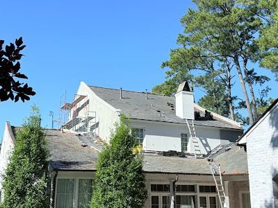 A white house with a roof undergoing repairs, featuring scaffolding and a ladder against the chimney under a blue sky.