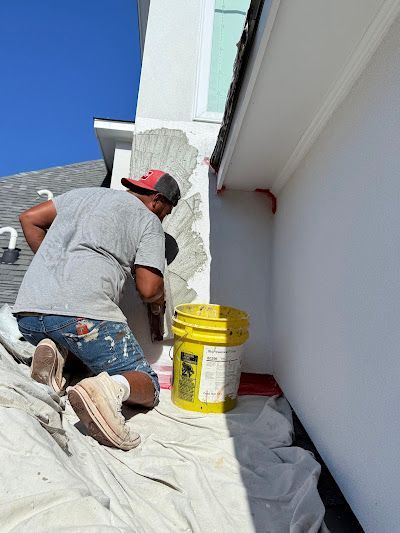 A person wearing a hat and work clothes patches a white wall corner with mortar, next to a yellow bucket on a drop cloth.
