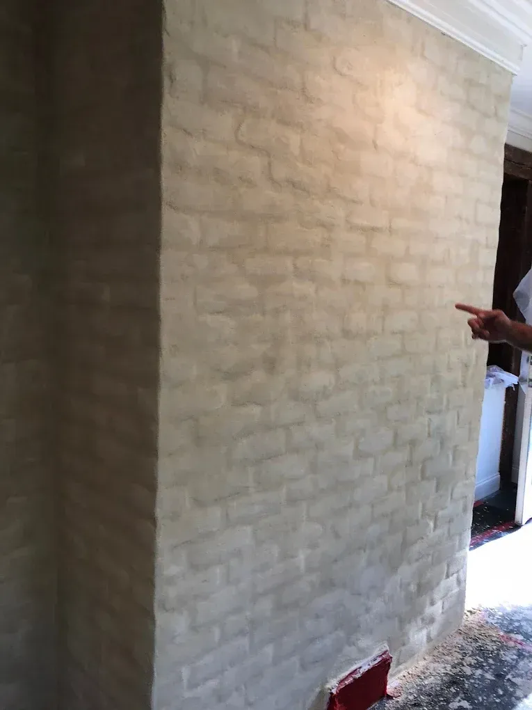 Bricked wall with light-colored coating. A person's hand points toward the wall. Setting is indoors.