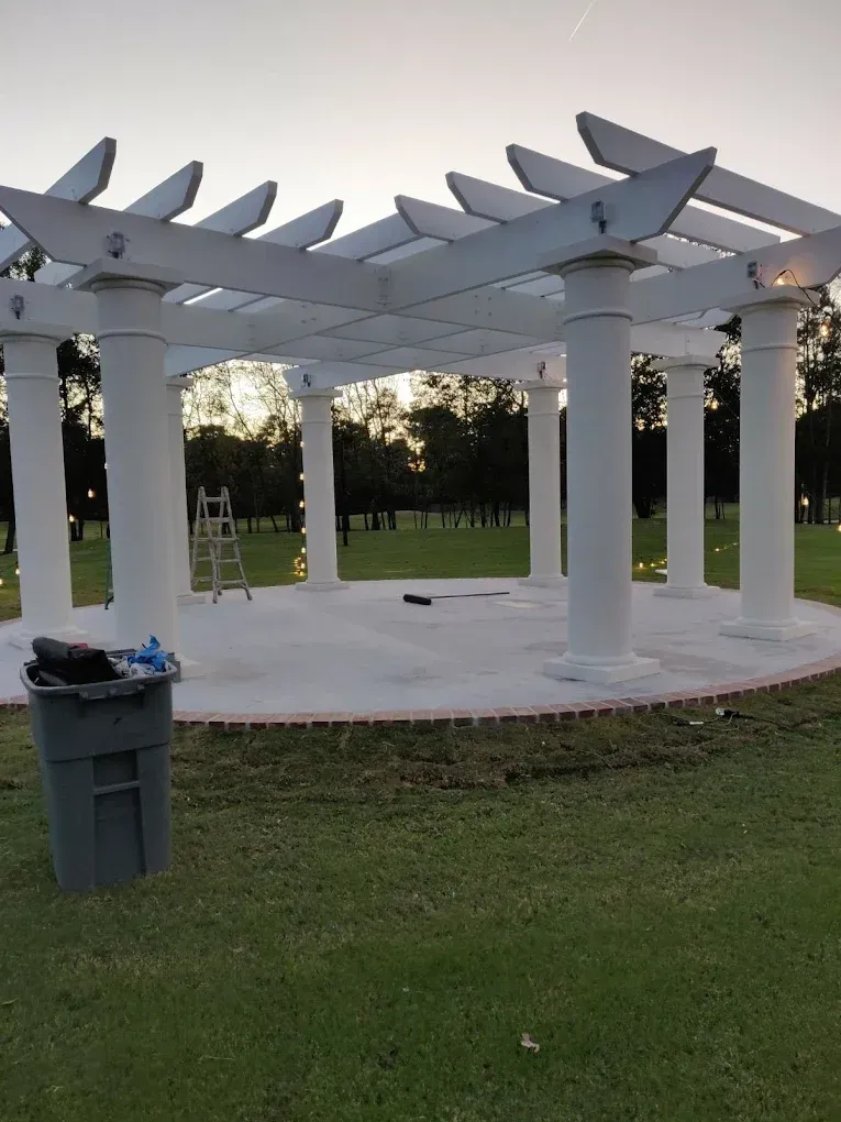 White gazebo with columns, on a concrete pad, surrounded by green grass. A trash can and ladder are nearby.