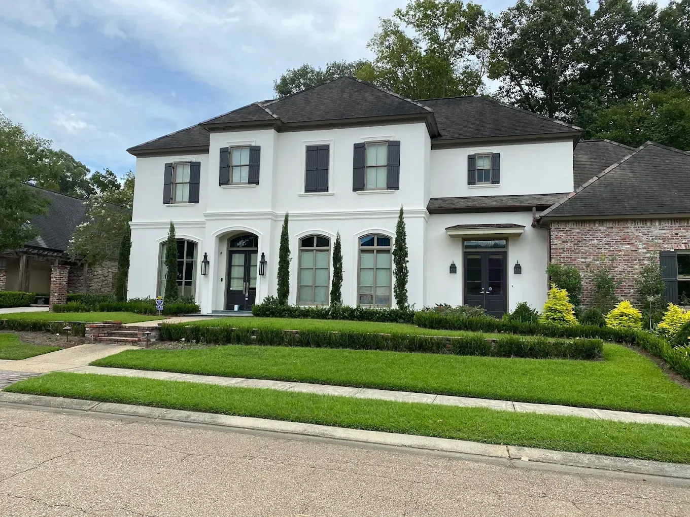 White two-story house with black shutters, green lawn, hedges, and trees on a sunny day.