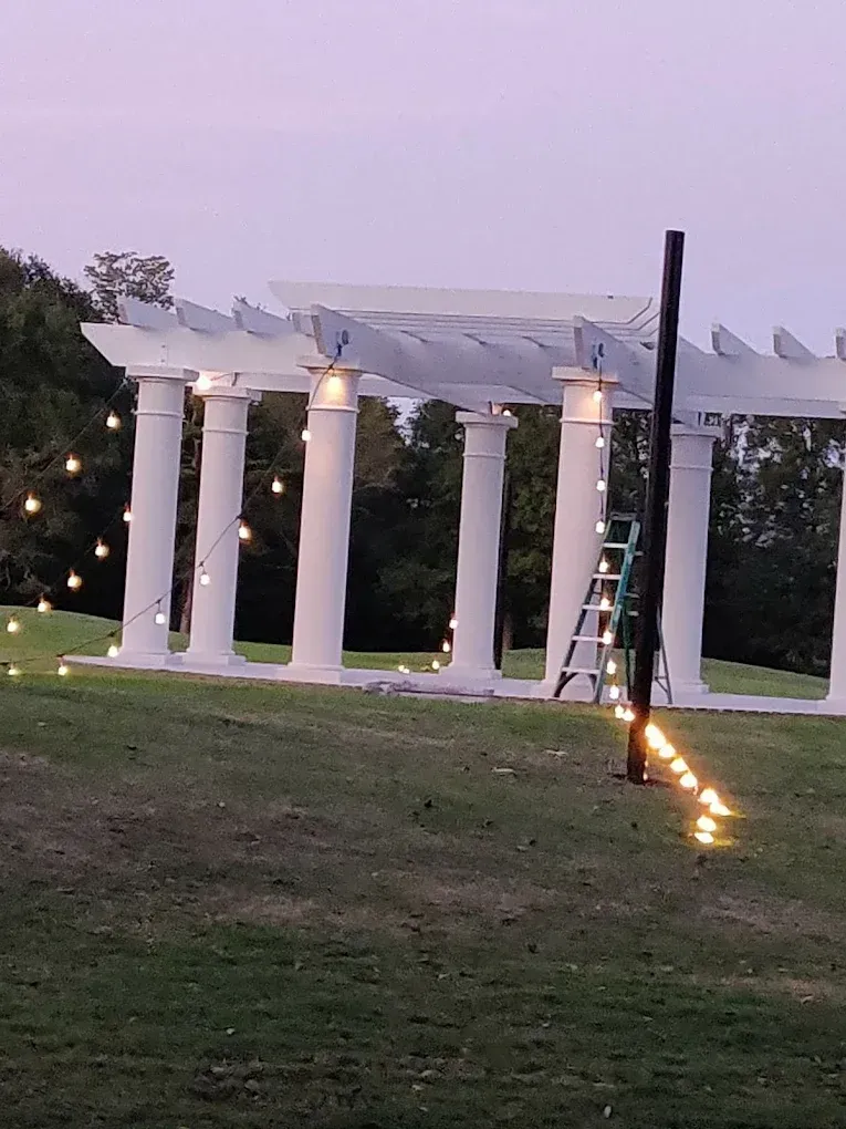 White columned pergola on a grassy hill, strung with lights at dusk. A ladder and pole stand nearby.