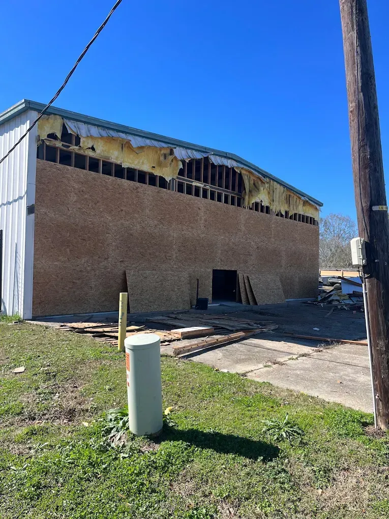 Brick building with damaged roof under a blue sky, construction materials on ground.