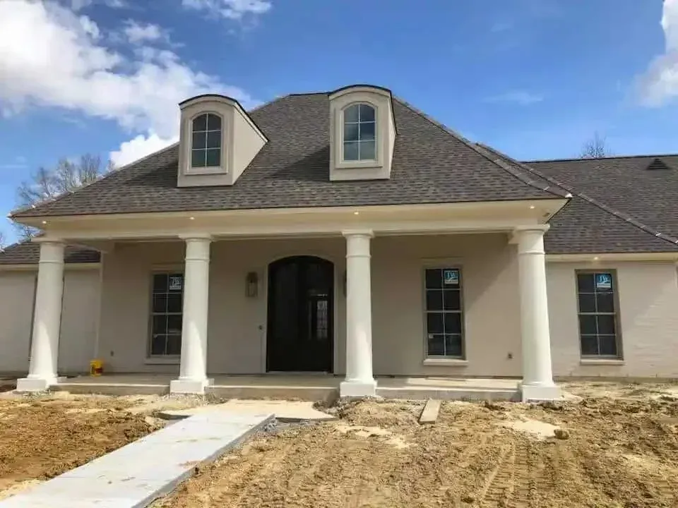 Beige house with porch, white pillars, dormer windows, dark roof, blue sky, construction site.