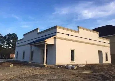 A light-colored, single-story building under construction with a flat roof and a small porch.