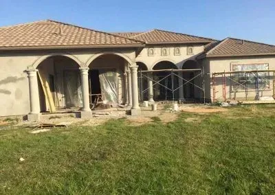 House under construction with columns, arches, and tan roof; green grass in foreground.