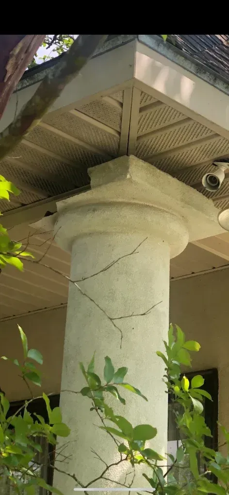 Concrete column with cracks, supporting a roof overhang. Green foliage in foreground.