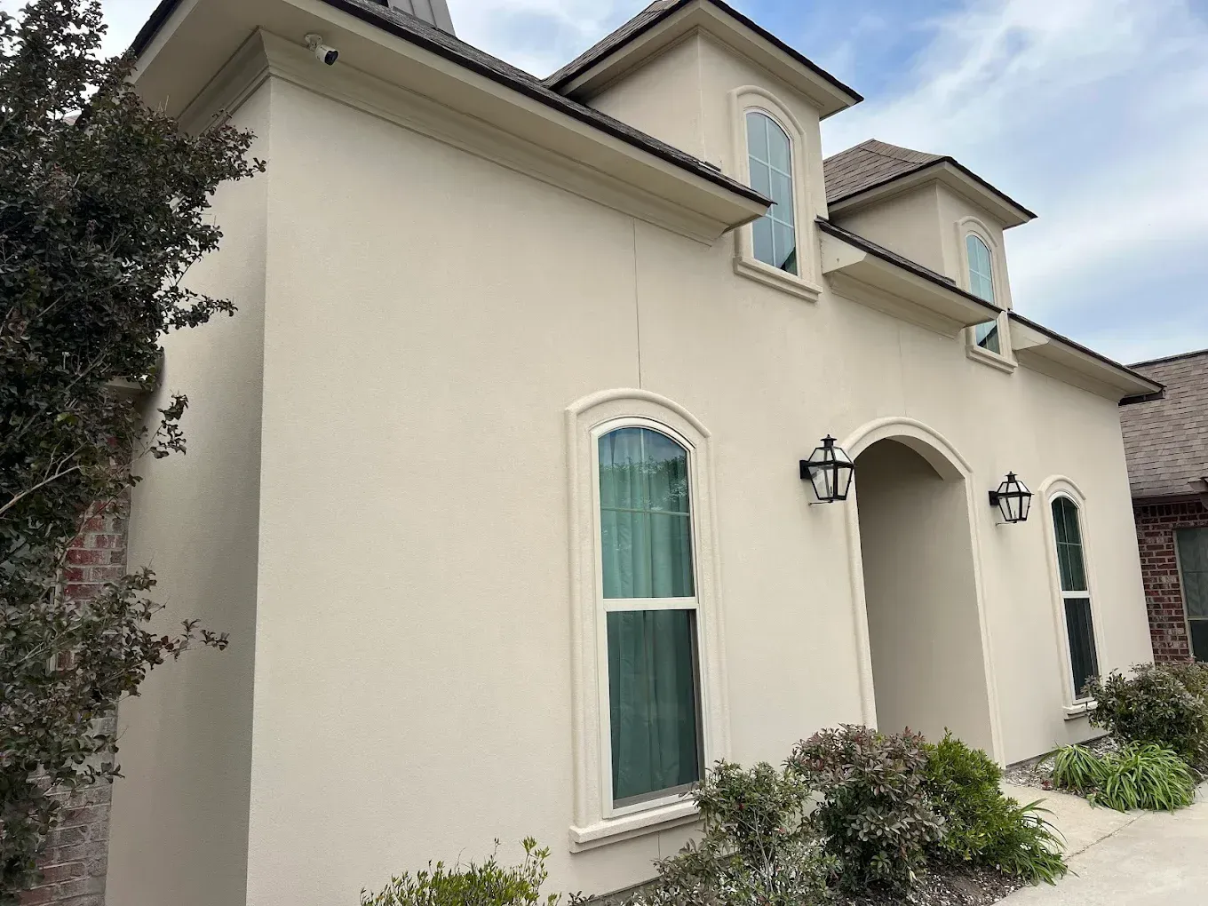 Beige stucco house with arched windows and entry, black sconces, and landscaping.