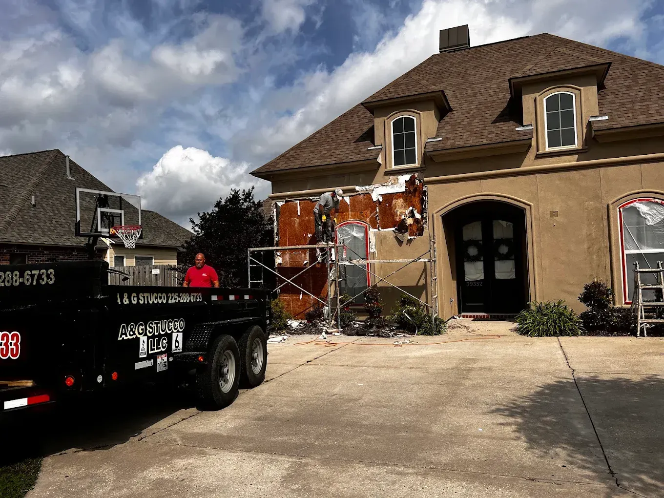 House with damaged exterior being repaired; worker stands by trailer.