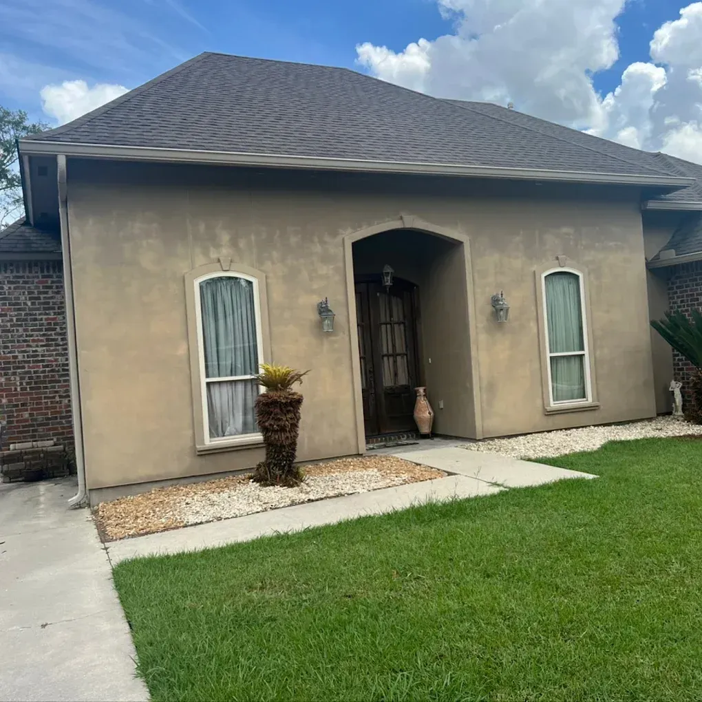 Beige stucco house with brown roof and arched entryway. Two windows flank the door; green lawn.