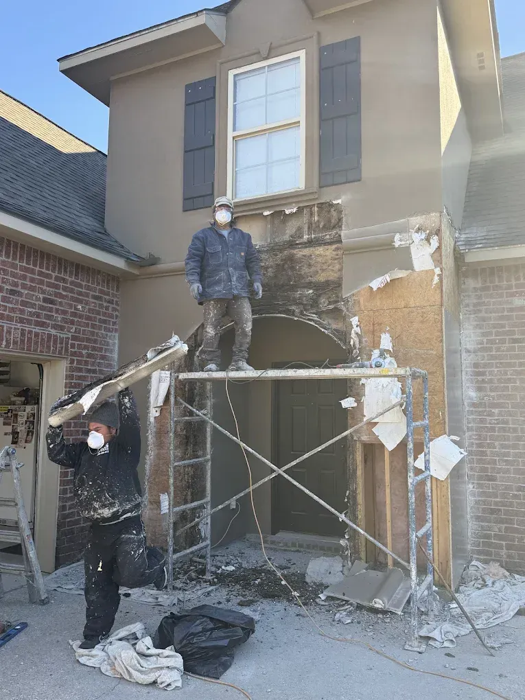 Two workers removing stucco from a house facade; one on scaffolding, one holding debris.