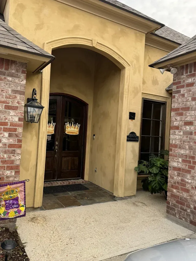 Exterior of a house entrance with dark wooden double doors, tan stucco walls, and brick accents.