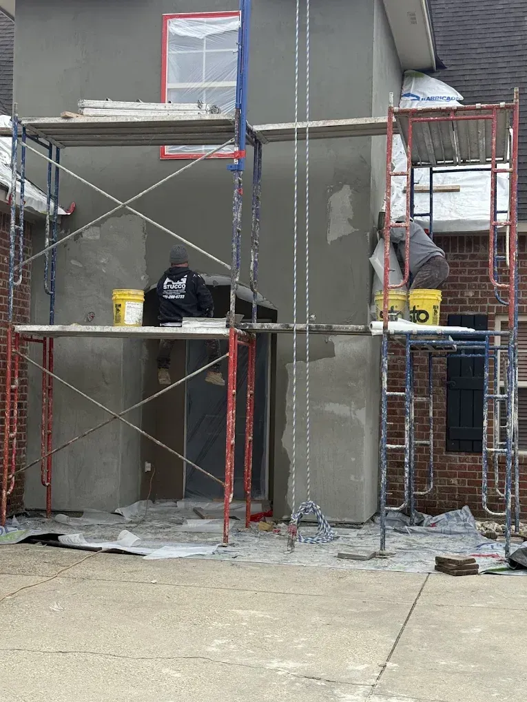 Two workers on scaffolding applying stucco to an exterior wall. Grey stucco on building, red and blue scaffolding.