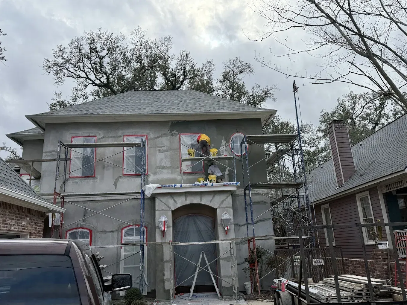 House exterior under construction, scaffolding, workers, gray stucco, red window trim, overcast sky.