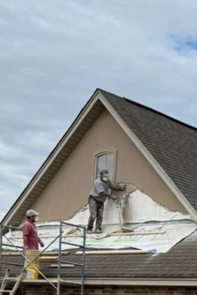 Workers on scaffolding repair stucco gable of a house with cloudy sky in the background.