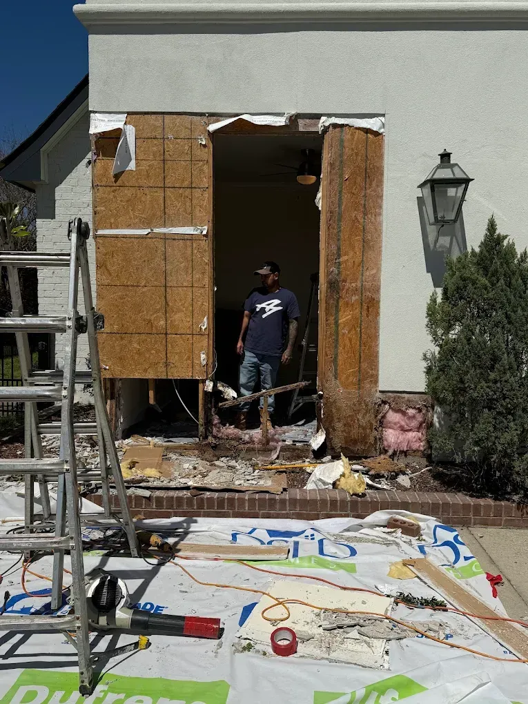 Construction site: man stands in doorway of a house undergoing renovation; tools and debris are scattered on the ground.
