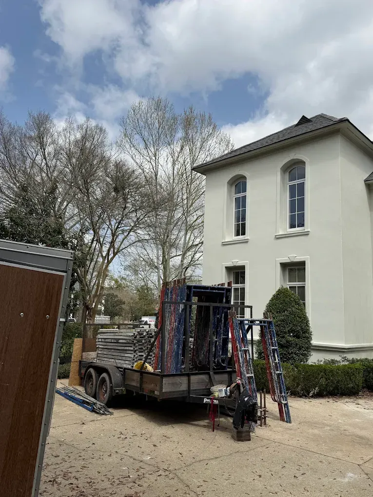 Trailer loaded with equipment parked near a two-story white building, cloudy sky above.