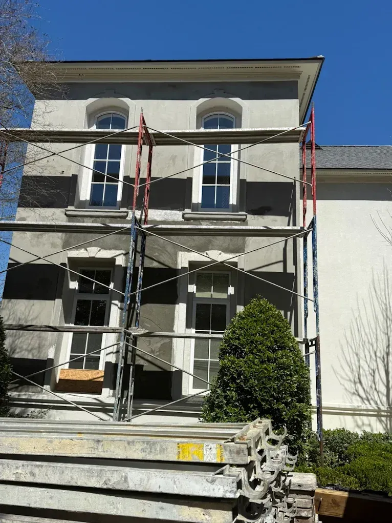 Building facade with scaffolding, undergoing renovations. Gray exterior, white window frames, bright blue sky.