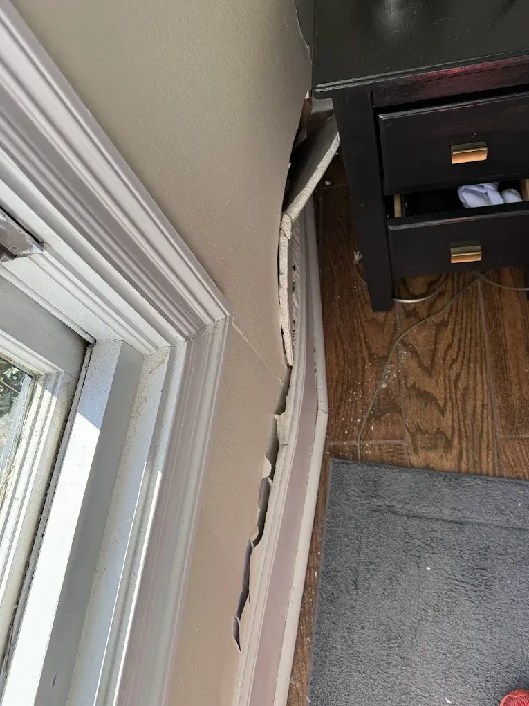 Damaged wall next to a nightstand and window frame; paint peeling.