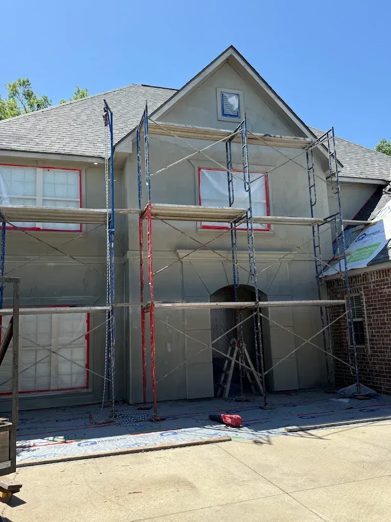 House exterior with scaffolding during stucco application, windows covered in red tape.