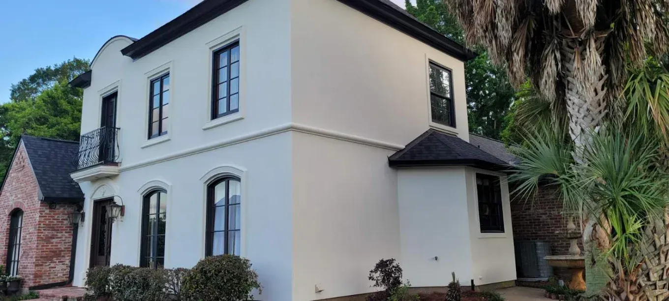 Cream-colored stucco house with black-framed windows, a dark roof, and brick side, set amongst trees.