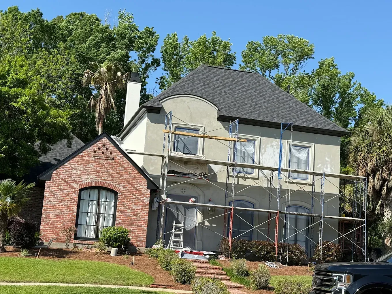 Two-story house under renovation, scaffolding, brick section, gray stucco, black roof, surrounded by green trees.