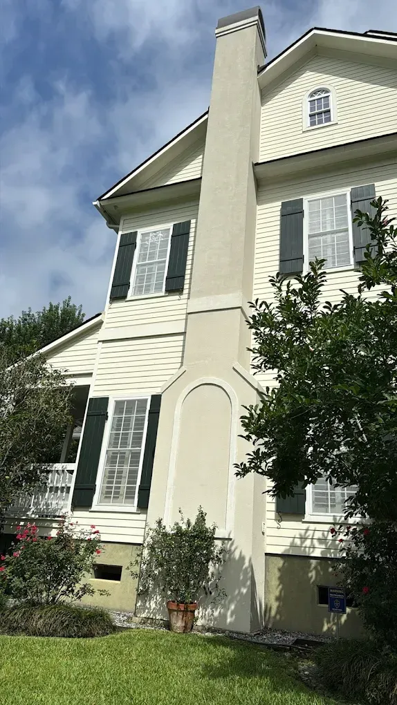 Beige and white two-story house with black shutters and a tall chimney against a cloudy sky.