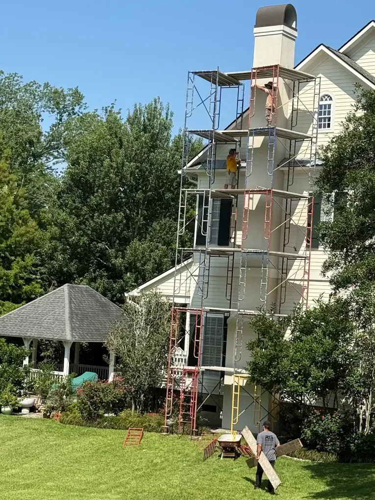 Workers on scaffolding next to a tall, cream-colored house. One carries a plank, the other is on top.