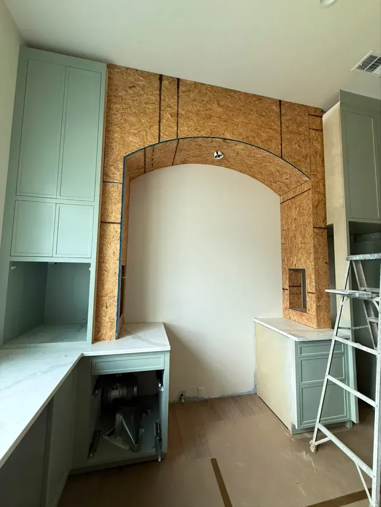 Kitchen under construction: arched OSB cutout, cabinets in blue-green, unfinished countertops, and ladder present.