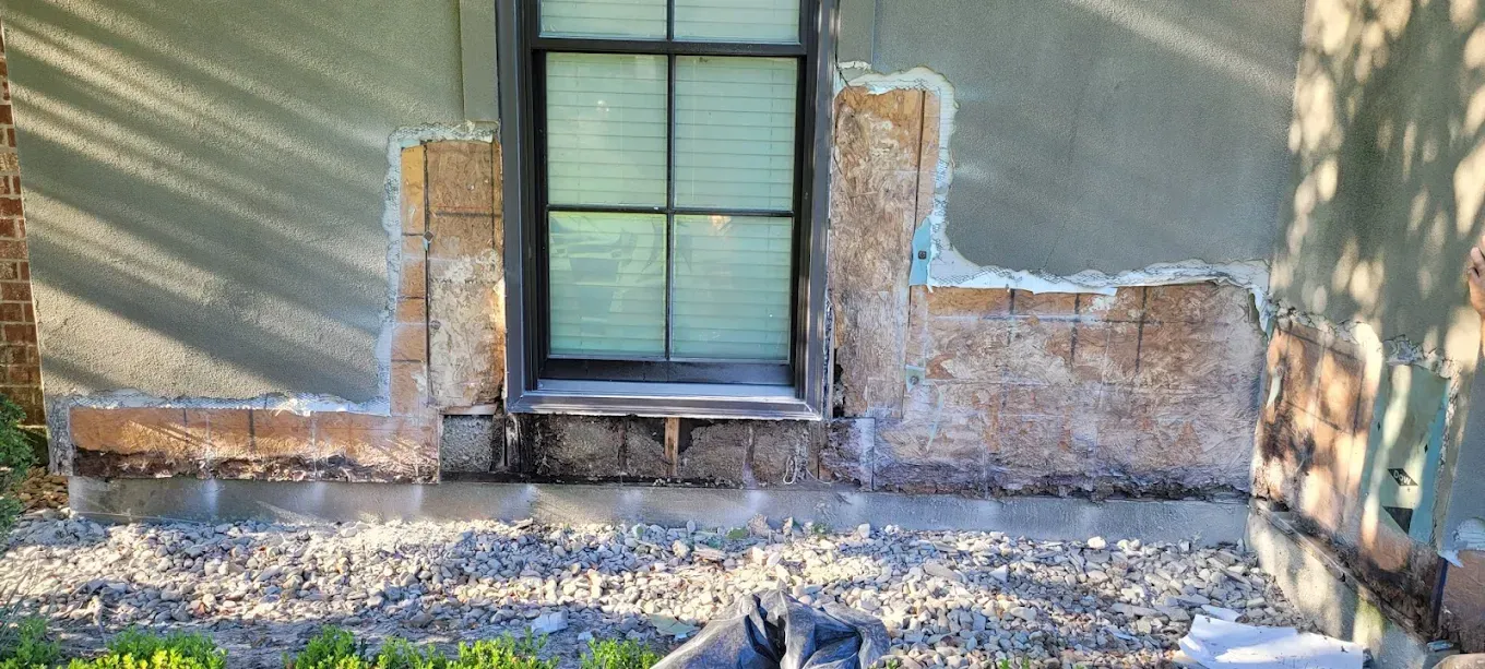 Exterior wall with window. Damaged stucco reveals brick or block construction. Ground is covered in gravel.