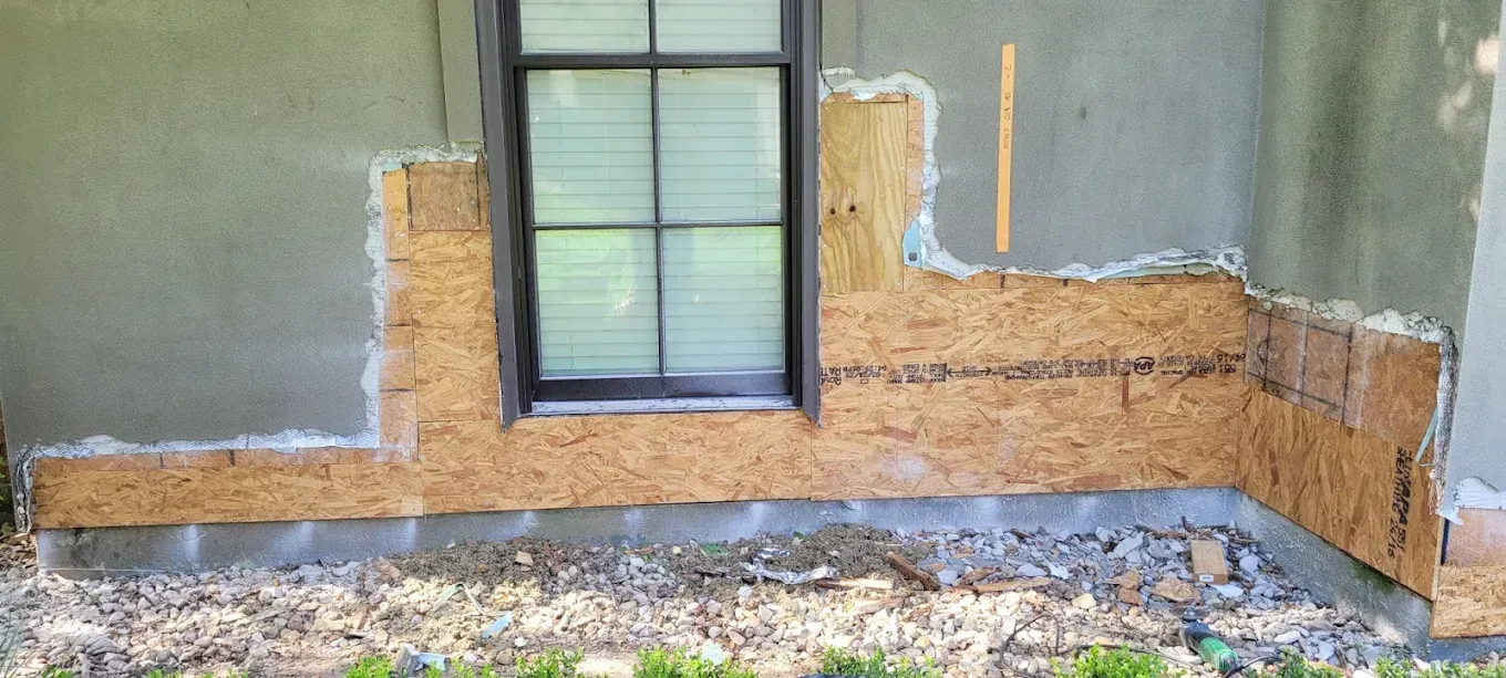 Exterior wall with exposed wood sheathing around a window, showing damaged stucco.