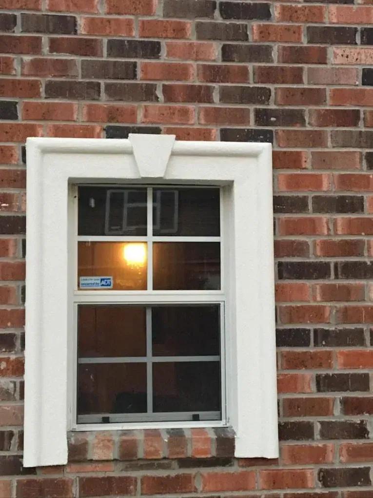 White framed window in a brick building reflects bright light, with a security sticker visible.